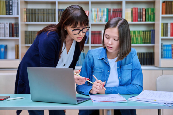 Young female university student with middle aged teacher mentor preparing for exam sitting at desk with laptop books inside library. Knowledge, education, youth, college university concept - Zdjęcie, obraz