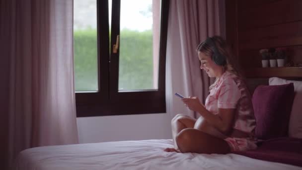 Boldog terhes nő otthon ül az ágyon mobilon. Vidám anya csatlakozik online, miközben pihentető és zenét hallgat fejhallgató. Az emberek fogalma a közösségi médiában és az anyaság. - Felvétel, videó