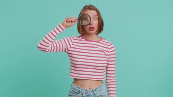 Investigator researcher scientist woman holding magnifying glass near face looking into camera with big zoomed funny eyes searching analysing. Young girl in crop top isolated on blue studio background - Footage, Video