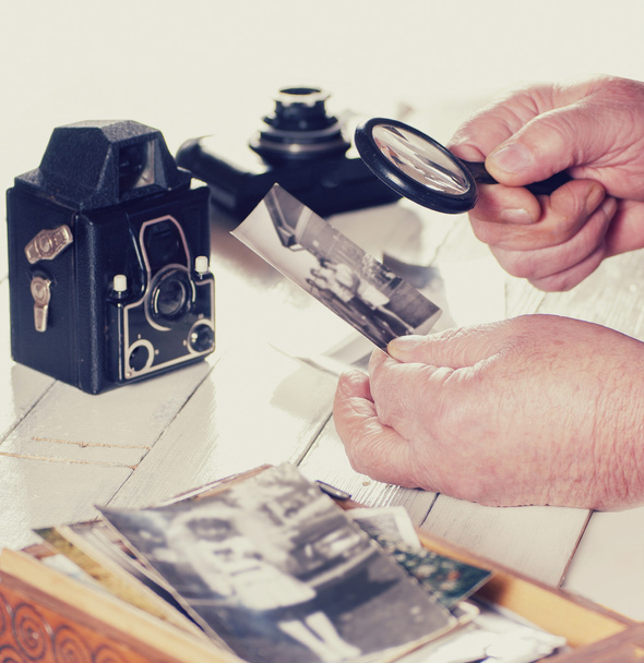 Παλιά άνθρωπος χέρια, προβολή φωτογραφιών με μεγεθυντικό φακό από το παρελθόν, ρετρό κάμερες στο τραπέζι, φιλτράρεται φωτογραφία - Φωτογραφία, εικόνα