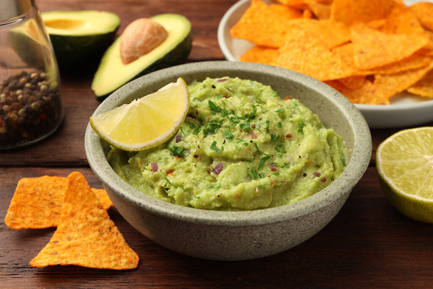Bowl of delicious guacamole, lime and nachos chips on wooden table, closeup - 写真・画像