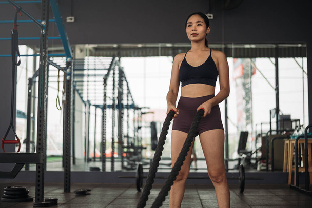 Erős ázsiai nő harci kötéllel tornázik a tornateremben. Sportoló nő visel sportruházat edzés szürke tornaterem háttér súly és súlyzó berendezések. Egészséges életmód. - Fotó, kép