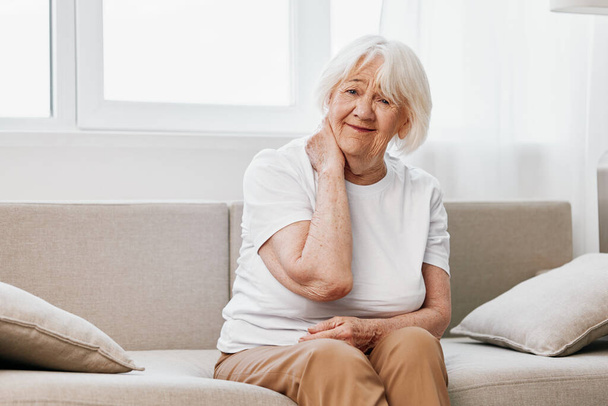 Mulher idosa dor intensa no pescoço sentada no sofá, problemas de saúde na velhice, má qualidade de vida. Foto de alta qualidade - Foto, Imagem
