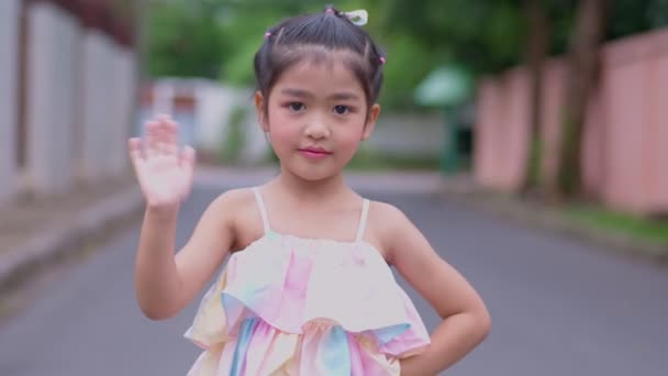 Retrato de uma menina asiática atraente sorrindo e olhar câmera e acenar com a mão, criança sorrindo feliz olhando para a câmera - close-up, ao ar livre. Natureza alegre conceito de lazer infantil. - Filmagem, Vídeo