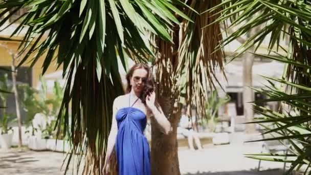 Egy kék ruhás lány sétál a pálmafalván egy fényes, napos napon. Kiváló minőségű 4k felvételek - Felvétel, videó
