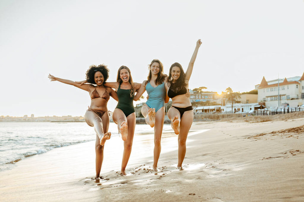 Sahilde güzel vakit geçiren dört mutlu kadın, sahilde yürürken kucaklaşıyorlar, genç kadınlar gülüyor ve okyanus kıyısında tatillerinin tadını çıkarıyorlar. - Fotoğraf, Görsel