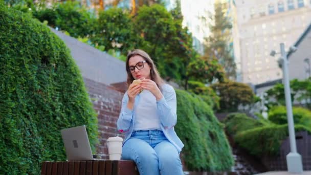 Mujer caucásica almorzando con un sándwich y café mientras está sentada en el parque con un portátil. Al aire libre. La freelancer trabaja con su portátil - Metraje, vídeo