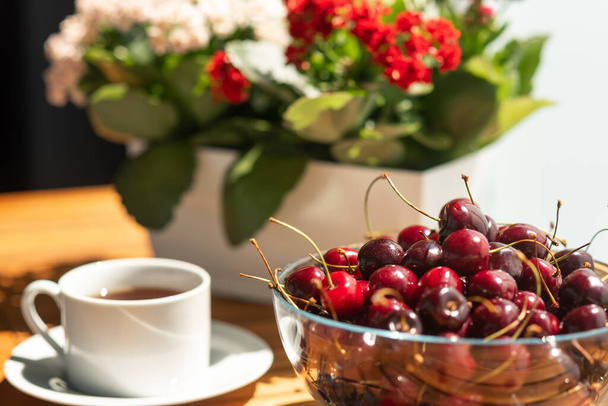 Cerises dans un grand bol en verre sur une table en bois. À côté d'une tasse de thé et de fleurs de kalanchoe dans une jardinière blanche. Journée ensoleillée d'été sur la terrasse - Photo, image