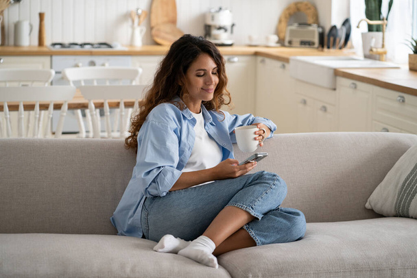 Beztroska młoda kobieta za pomocą smartfona, odpoczynku z filiżanką herbaty w przytulnym domu, zrelaksowany kobieta siedzi na kanapie ciesząc się ulubionym gorącym napojem, przewijanie sieci społecznych na telefon komórkowy. Koncepcja wolnego życia - Zdjęcie, obraz