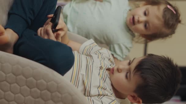Dos niños jugando videojuegos en smartphone, descansando en sillones en casa solos. Curioso inteligente gen Z hermano y hermana - Imágenes, Vídeo