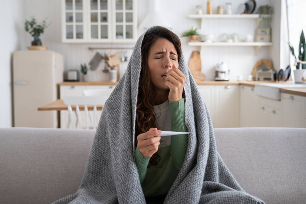 Женщина, сидящая дома на диване, страдает насморком, чихает. Больная женщина, завернутая в одеяло и удерживающая температуру термометра, страдает от симптомов простуды или гриппа. Зимние вирусы - Фото, изображение