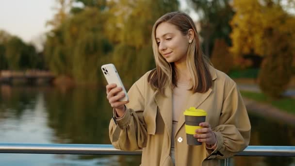 Mooie vrouwelijke influencer staan in het park met behulp van haar smartphone voor sociale media, sms 'en, het vasthouden van de koffie. Blanke dame buiten Scrolling Mobile, chatten Browsing Online - Video
