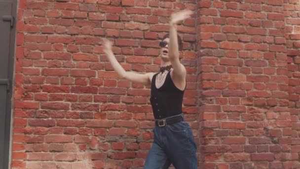 Feminine boy in black shades, choker and earrings voguing against red brick wall outdoors - Footage, Video