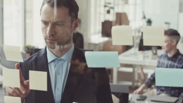 Serieuze blanke zakenman in formalwear peeling plakkerige notitie met taken om te doen van glas raam tijdens het werken in IT-bedrijf - Video