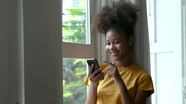 スマートフォンを使って自宅でメッセージを閲覧するスタイリッシュな黒人女性リラックスしたライフスタイルを楽しむ窓の外を見るアフリカ系アメリカ人女性携帯電話でテキストメッセージ. - 映像、動画