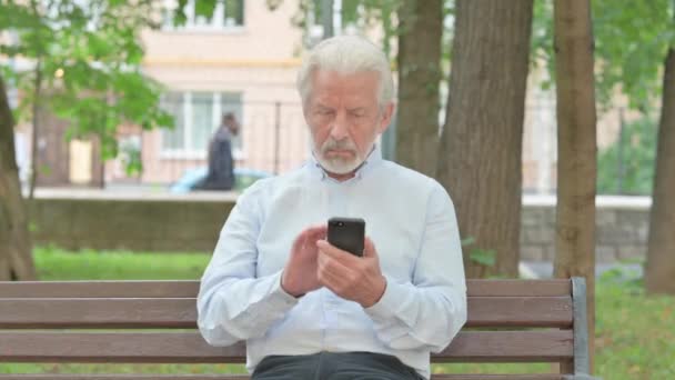 Vecchio anziano che naviga su Internet Smartphone mentre è seduto all'aperto su una panchina - Filmati, video