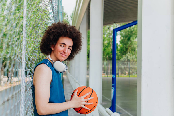 Portrét dospívajícího chlapce s kudrnatými vlasy a sportovním oblečením a sluchátky držícího basketbalový míček v městském kurtu - Fotografie, Obrázek