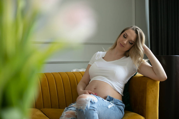 Удивительная молодая беременная женщина сидит на жёлтом диване, прислонившись головой к руке и нежно касаясь голого живота. Блондинка в белой футболке и порванных джинсах отдыхает дома, цветы на переднем плане. - Фото, изображение