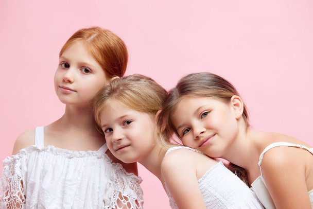 Three beautiful, cute little girls, children in ponytails, in white clothes looking at camera against pink studio background. Concept of skincare, childhood, cosmetology, health, beauty, wellness, ad - Photo, image