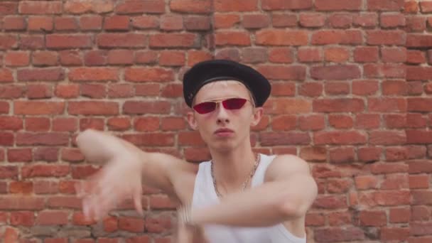 Stylischer Junge mit weißem Tank-Top, Baskenmütze und Vintage-Schattierungen, der vor roter Backsteinwand im Freien tanzt - Filmmaterial, Video