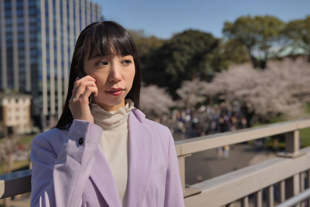 Un retrato de una mujer japonesa llamando a un teléfono inteligente detrás de la flor de cerezo. Foto de alta calidad. Distrito de Chiyoda Tokio Japón 03.30.2023 - Foto, Imagen