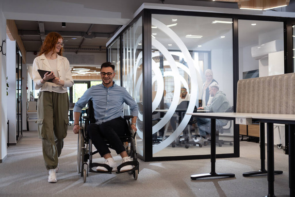 Young business colleagues, collaborative business colleagues, including a person in a wheelchair, walk past a modern glass office corridor, illustrating diversity, teamwork and empowerment in the - Photo, Image