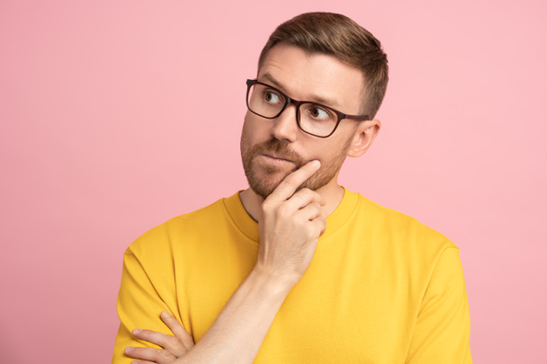 Thoughtful puzzled American man look away holding hand on chin. Pensive student or entrepreneur in eyeglasses ponder making decision of investment looking at empty copy space over studio pink wall - Photo, Image