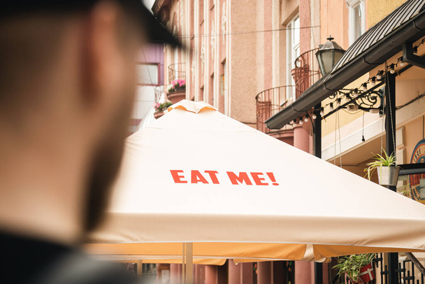 Άνδρας τουρίστας ψάχνει σε μια ομπρέλα βεράντα εστιατόριο με επιγραφή Τρώτε μου από ένα καφέ δρόμο, έννοια του street food, ένα σνακ, ενώ ταξιδεύετε σε μια ξένη πόλη. - Φωτογραφία, εικόνα