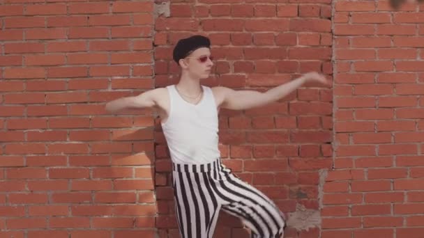 Ragazzo femminile che indossa tonalità vintage, pantaloni a righe in bianco e nero e basco danza voga contro muro di mattoni rossi all'aperto - Filmati, video