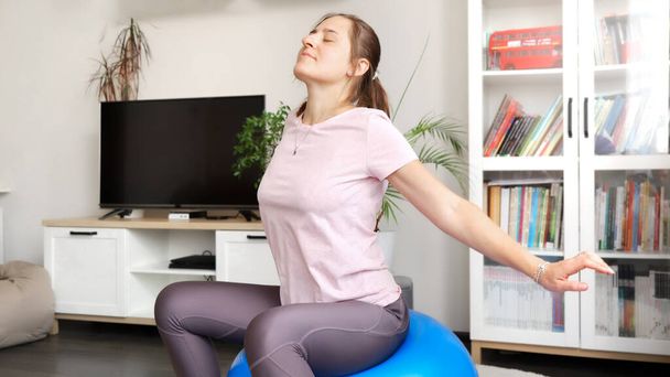 Νεαρή χαλαρή γυναίκα με ρούχα γυμναστικής κάθεται σε fit μπάλα και κάνει ασκήσεις αναπνοής. - Φωτογραφία, εικόνα