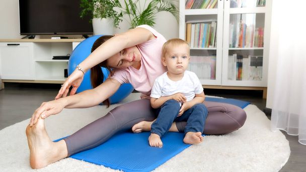 Cute baby boy sitting on fitness mat and looking at his mother stretching legs before sports training. Concept of healthcare, sports and yoga at home - Photo, Image