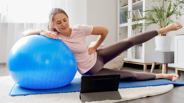 Νεαρή γυναίκα βλέποντας σε απευθείας σύνδεση μάθημα φυσικής κατάστασης βίντεο και κάνει ασκήσεις με fitball. Έννοια της υγειονομικής περίθαλψης, του αθλητισμού και της γιόγκα στο σπίτι - Φωτογραφία, εικόνα