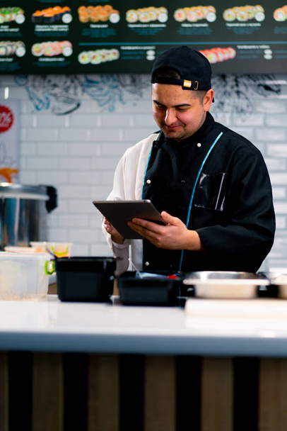 Przemyślane Sushi Chef z tabletką w rękach w pobliżu stołu kuchennego, na którym produkty do rolek są układane tobiko roe krewetki łososiowe krewetki - Zdjęcie, obraz
