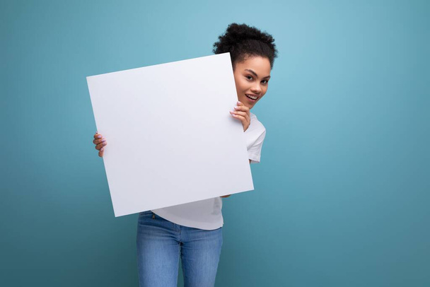 jeune femme brune avec des cheveux afro dans un débardeur blanc tenant une feuille de tableau blanc avec espace pour l'information. - Photo, image