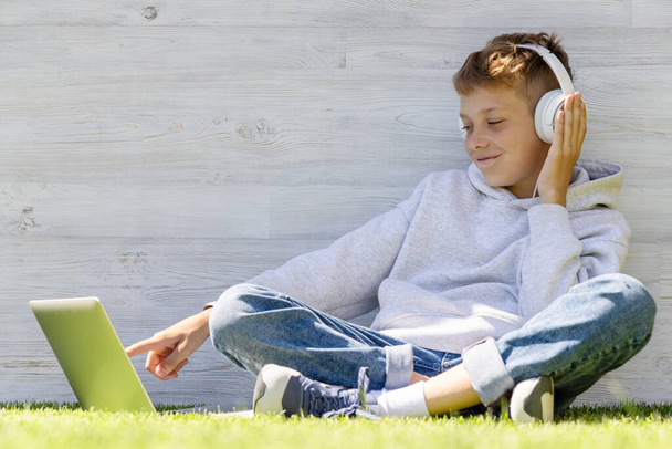 Poika käyttää kannettavaa tietokonetta ja kuulokkeita ulkona. Koulutuksen tai viihteen käsite - Valokuva, kuva