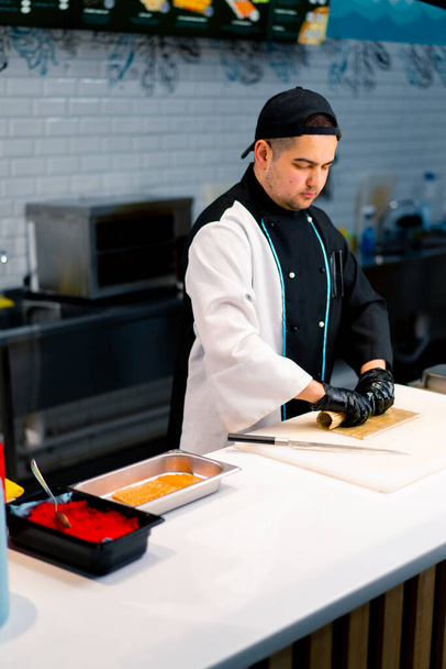 Suşi şefi, beyaz mutfak tahtasının üzerinde kurutulmuş somon krem peyniri ve salatalık ile Philadelphia usulü rulo hazırlıyor. - Fotoğraf, Görsel