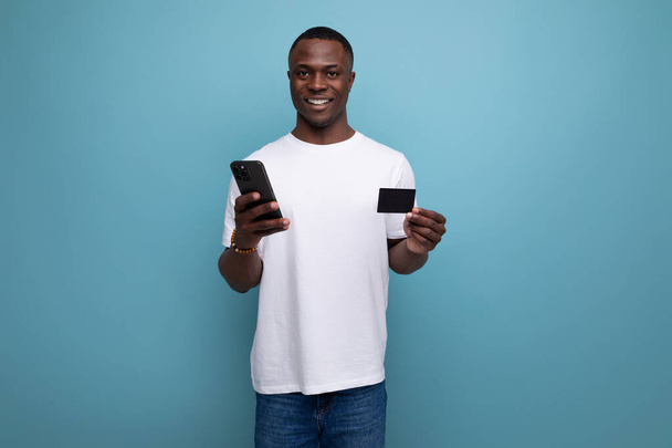 Beyaz tişörtlü genç Afrikalı olumlu bir adam akıllı telefon ve kartla internetten satın alımları ödüyor.. - Fotoğraf, Görsel