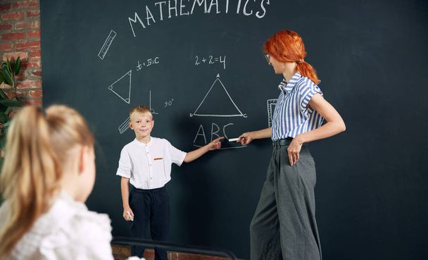 Young woman, teacher standing by blackboard with little boy, child and teaching math at school. Solving geometry tasks. Concept of school, education, childhood, knowledge, lifestyle - Φωτογραφία, εικόνα