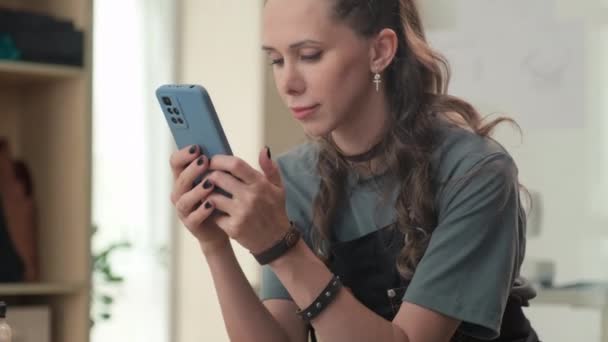 Μεσαία φωτογραφία του δέρματος craftswoman χρησιμοποιώντας smartphone για να ελέγξετε τα μηνύματα από τους πελάτες στο εργαστήριο - Πλάνα, βίντεο