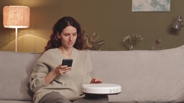 Nuori brunette Valkoihoinen nainen kodikas pusero istuu sohvalla olohuoneessa yhdistää uuden robotti tyhjiö puhelimeensa - Materiaali, video