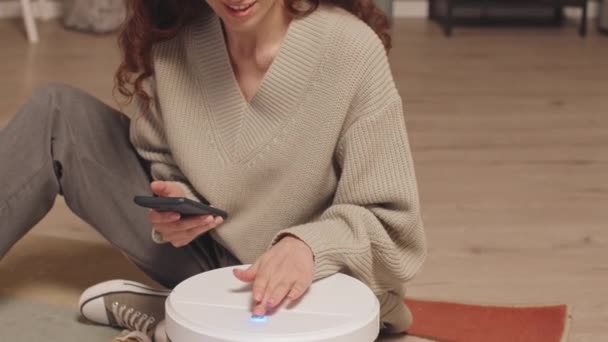 Junge brünette kaukasische Frau mit ihrem Telefon beim Einrichten neuer Staubsaugroboter, sitzt auf dem Boden im gemütlichen Wohnzimmer - Filmmaterial, Video