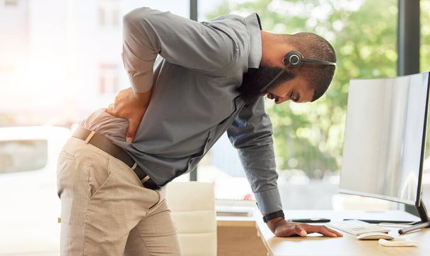 Call center, back pain and a black man consultant in his office, standing while holding is spine. Injury, customer service or support and a young male employee with muscle strain in a crm workplace. - Photo, Image