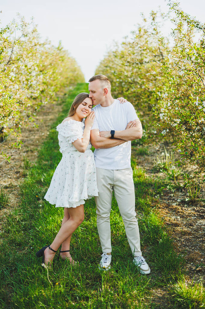 A beautiful young couple expecting a pregnancy in a romantic place, a spring blossoming apple orchard. Happy joyful couple enjoying each other while walking in the garden. A man holds a womans hand - Photo, image