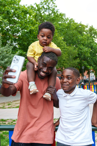 Beaming African dad snaps a vibrant selfie with his kids in a colorful green park; baby perched on shoulders, pure happiness." - Photo, Image