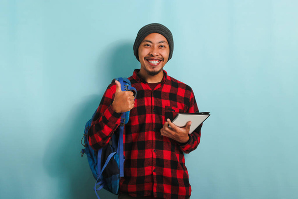 Een lachende jonge Aziatische student met een muts en een rood geruite flanellen hemd met rugzak geeft een duim naar de camera terwijl hij tegen een blauwe achtergrond staat - Foto, afbeelding