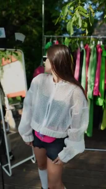 Nuori ja värikäs tyttö kokeilee takkia kesämarkkinoilla tanssiessaan. Laadukas FullHD kuvamateriaalia - Materiaali, video