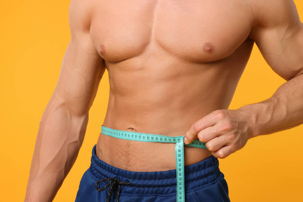 Athletic man measuring waist with tape on orange background, closeup. Weight loss concept - Foto, imagen