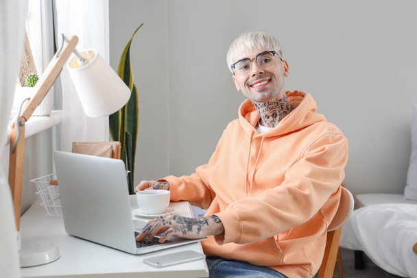 Joven hombre tatuado en gafas con portátil y taza de café en casa - Foto, Imagen
