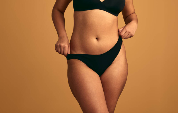 Crop anonymous plus size female model in black lingerie fixing panties standing against beige background - Foto, Imagen