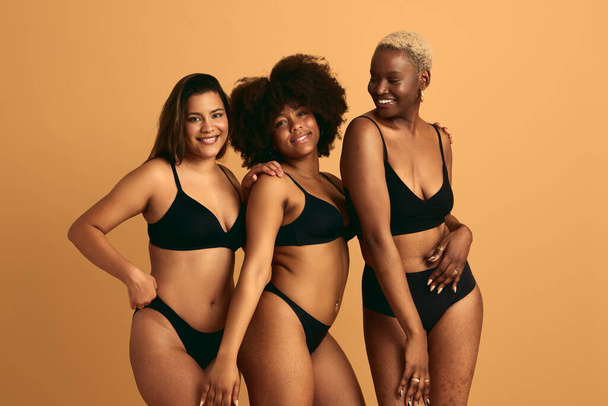 Group of self assured young multiracial women in black lingerie smiling while standing together against beige background - Foto, Bild